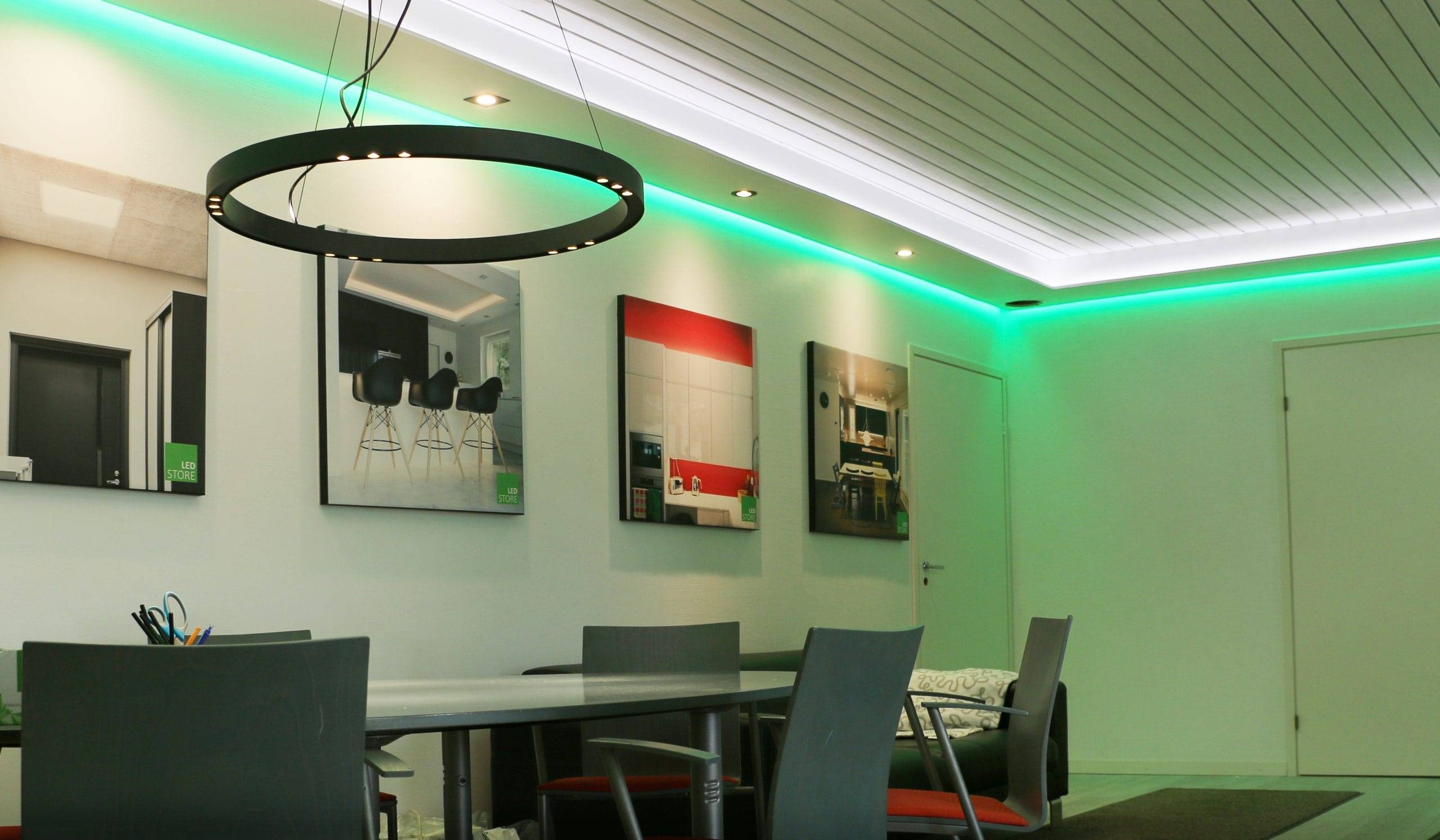Una sala moderna con una luz LED circular negra, sillas grises alrededor de una mesa larga, arte enmarcado en la pared e iluminación en el techo con reflejos verdes para crear un ambiente elegante.