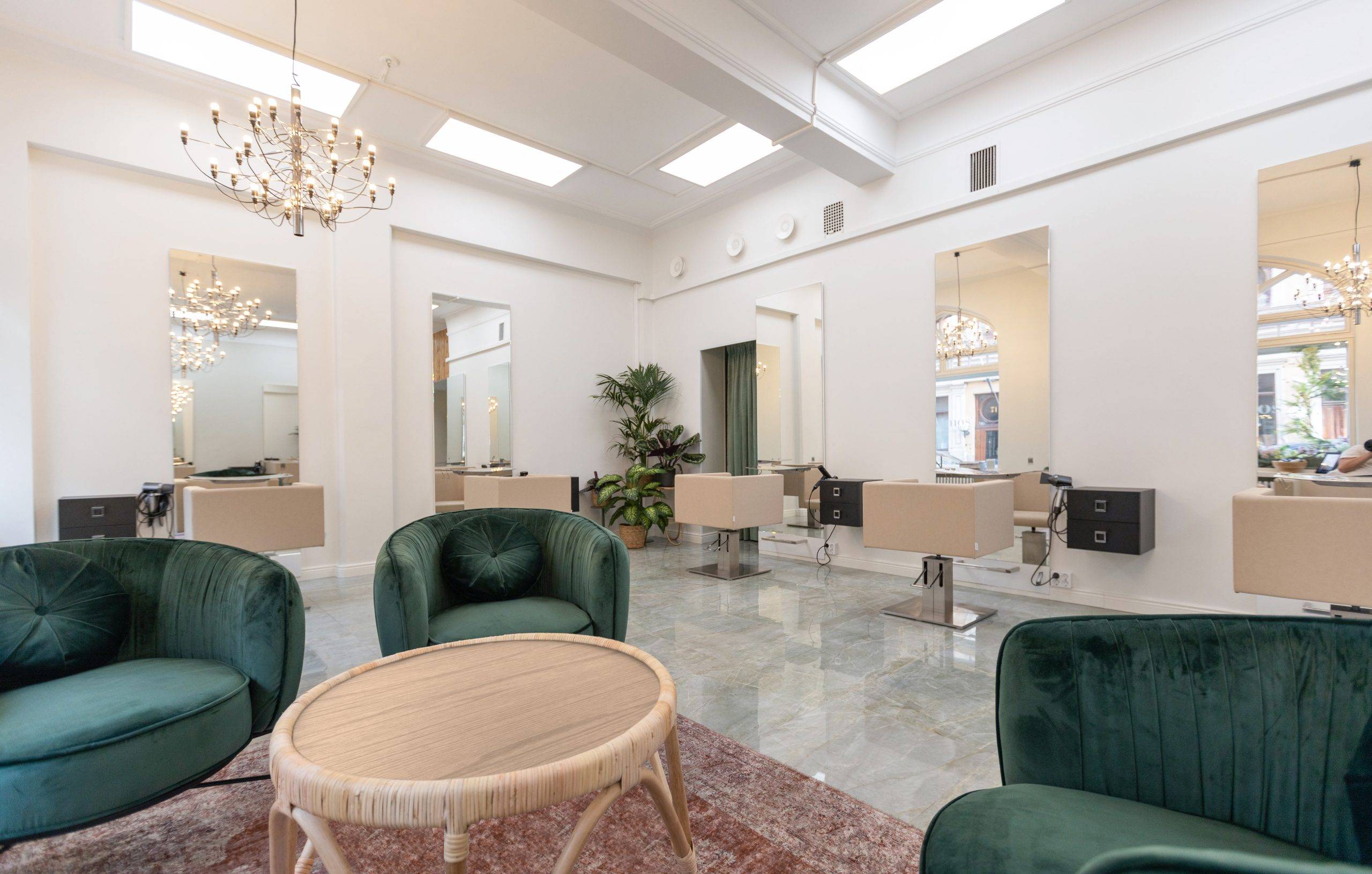 Un interior de salón moderno y luminoso con sillas de terciopelo verde que rodean una mesa redonda con una alfombra roja. Los puestos de trabajo del salón, con espejos y sillas beige, se alinean en las paredes. Están iluminados por lámparas de araña LED montadas en el techo. Los grandes ventanales dejan entrar la luz natural, mientras que la vegetación realza el ambiente del espacio. En este tipo de entorno, las soluciones de iluminación LED de alta calidad de LedStore.fi son ideales, ya que crean el equilibrio perfecto de luz y ahorro energético sin comprometer la elegancia.