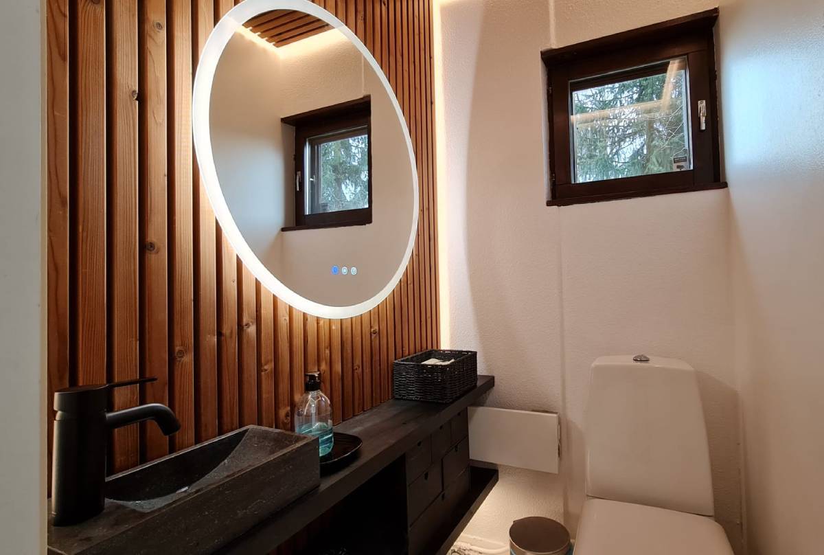 Un baño moderno con iluminación LED suministrada por LedStore.fi incluye un espejo LED circular encima de una pared de paneles de madera. Debajo del espejo hay una encimera negra con un lavabo rectangular y un grifo y dosificador de jabón negros. Encima del asiento del inodoro hay una pequeña ventana y cerca una práctica solución de almacenaje.