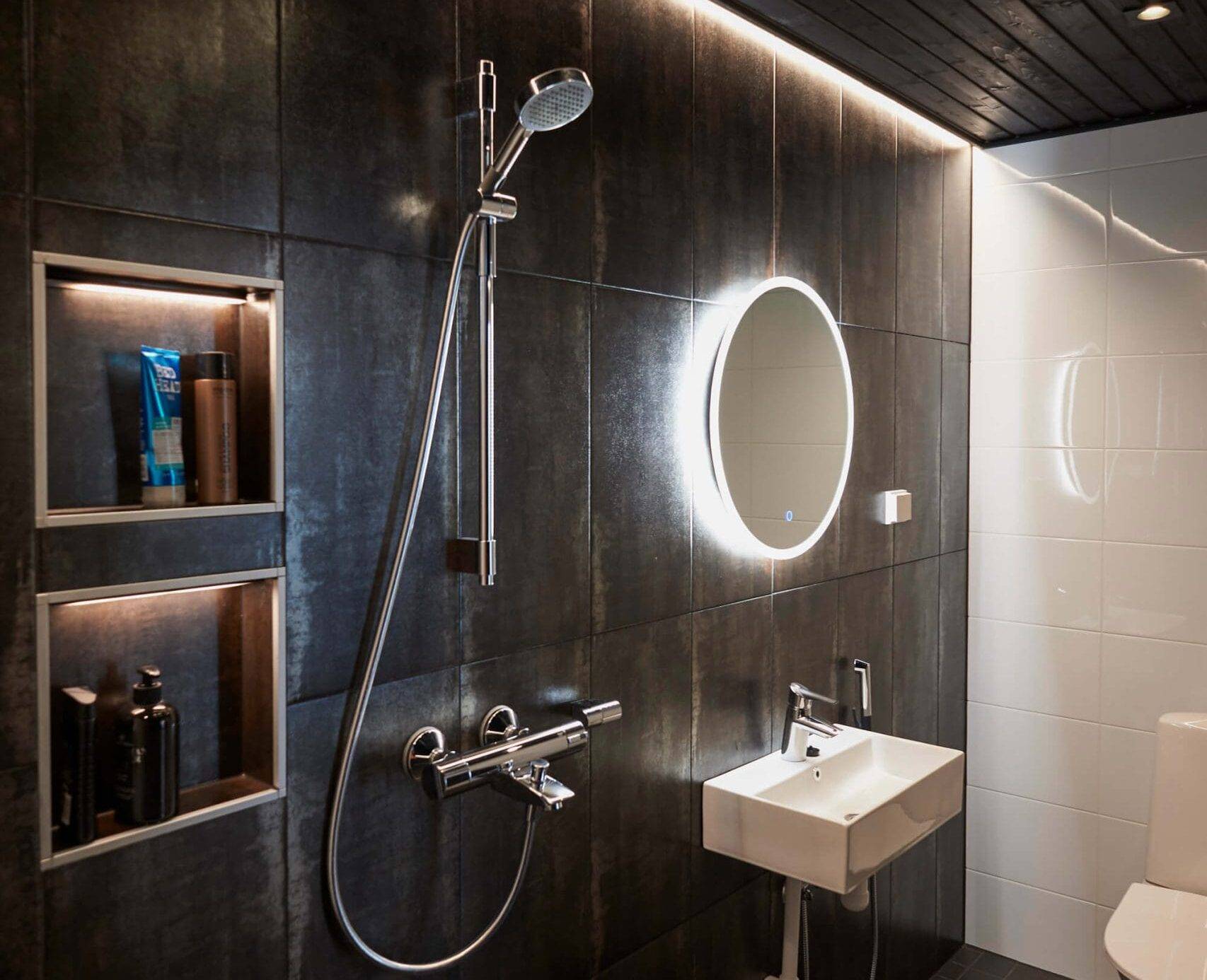 Elegante cuarto de baño con azulejos oscuros, espejo retroiluminado con LED sobre el lavabo, ducha, estanterías empotradas y techo negro. Un inodoro moderno completa el aspecto elegante.