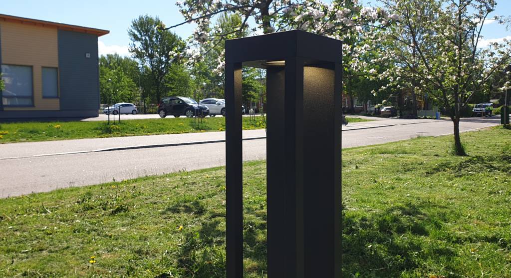 Una moderna farola negra con luz LED junto a la carretera; árboles en flor, coches aparcados y un edificio con grandes ventanales al fondo, bajo un cielo soleado.