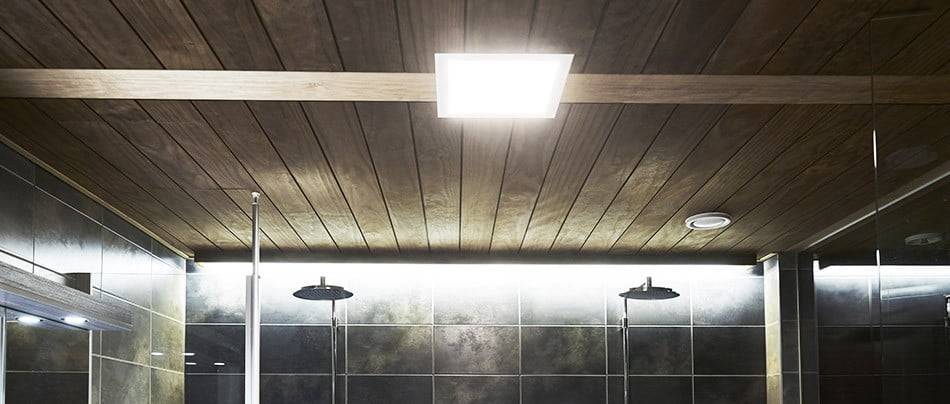 Moderno cuarto de baño con techo de madera, luminaria LED cuadrada, paredes de azulejos oscuros, dos rociadores de lluvia y un ambiente cálido con suave iluminación LED.