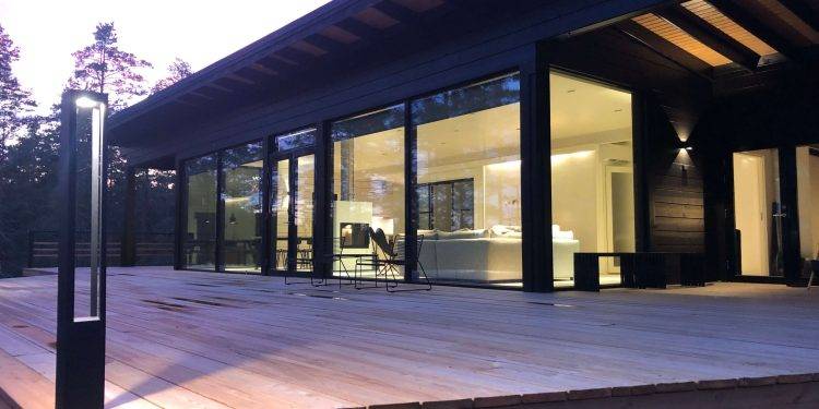 Casa moderna con grandes ventanales de cristal y espaciosa terraza de madera, rodeada de árboles. Los interiores están muy iluminados y muestran un diseño minimalista en tonos pintados a mano. Las luces LED exteriores de LedStore.fi añaden un cálido resplandor al crepúsculo vespertino.