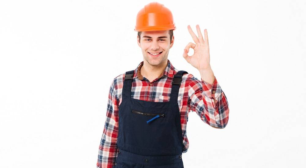 Retrato de un alegre joven constructor que muestra el gesto del pulgar hacia arriba mientras está de pie y mira a la cámara, aislado sobre fondo blanco.