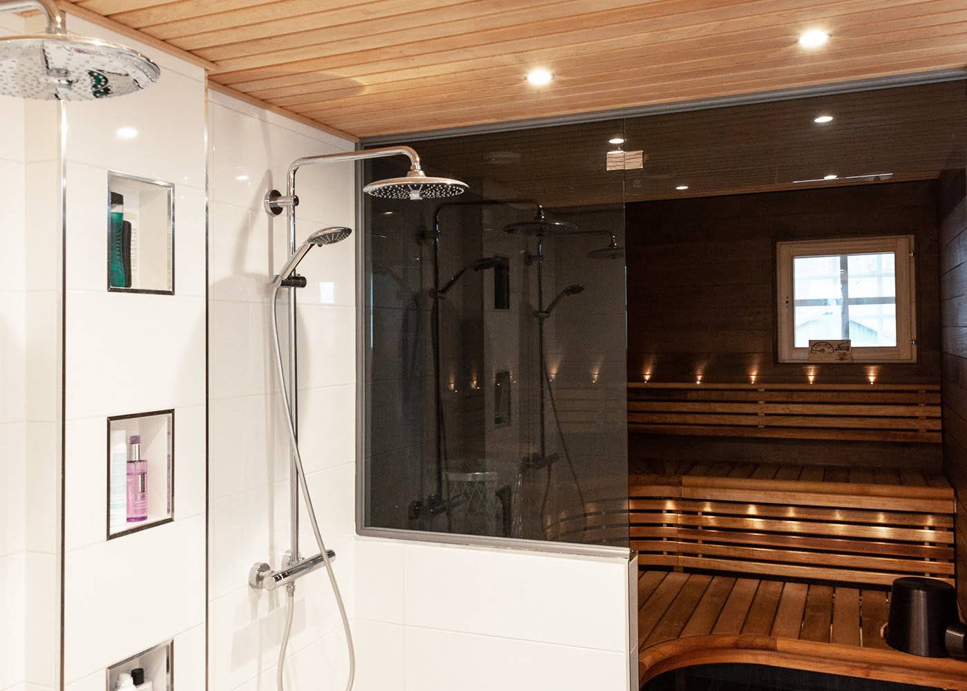 Moderno cuarto de baño con dos duchas efecto lluvia, pared de cristal, estante incorporado para artículos de aseo, sauna de madera contigua, iluminación atmosférica en las ventanas e iluminación LED.