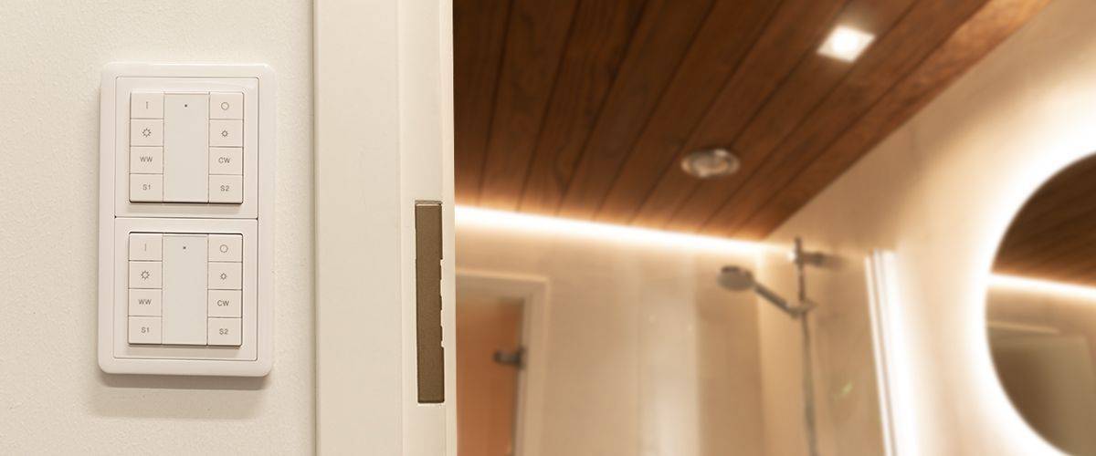 Cuarto de baño con paneles de madera en el techo e iluminación LED suave. También hay una práctica ducha a ras de suelo con cabezal de ducha tipo lluvia. En una pared cercana hay dos modernos paneles de interruptores de luz, controlados por teclados. El elegante conjunto está coronado por un espejo circular iluminado con LED.